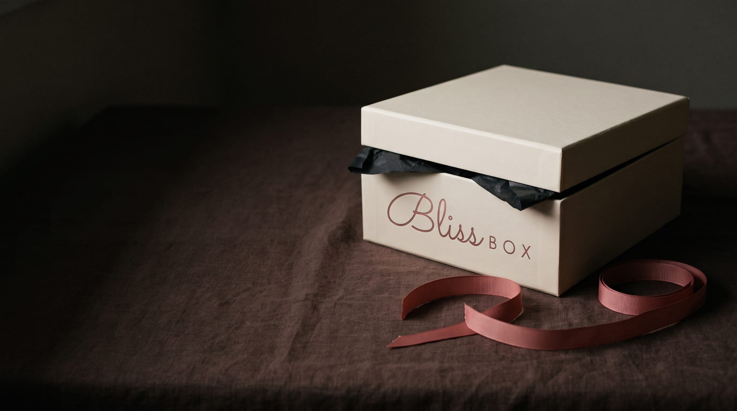 A Bliss Box gift box, lid ajar over black tissue, with a loose pink ribbon on dark linen